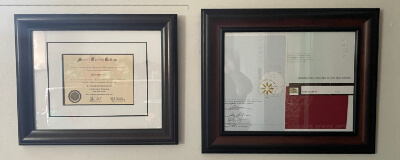 Two framed diplomas in dark brown frames hung on a white wall.