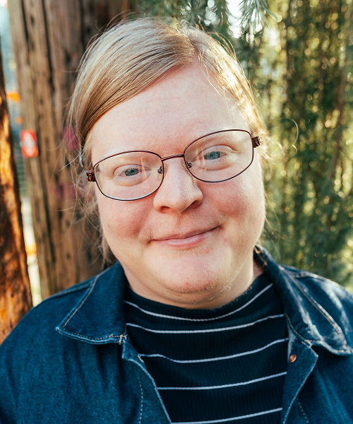 Rory Hackney, a white person in a blue jacket, is smiling in front of some trees.